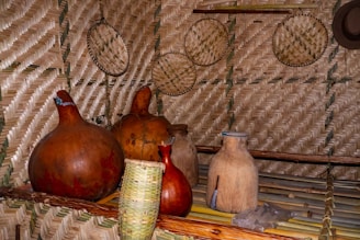 A rustic interior is adorned with intricately woven straw mats on the walls and several large, traditional earthenware pots and gourds on wooden shelves. Basketry and a hat are mounted as decorations.