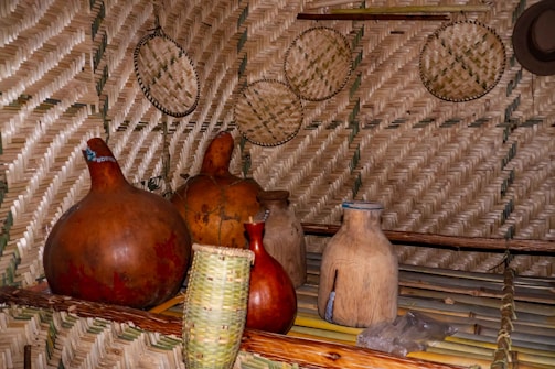 A rustic interior is adorned with intricately woven straw mats on the walls and several large, traditional earthenware pots and gourds on wooden shelves. Basketry and a hat are mounted as decorations.