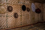 A wall made of woven bamboo with several hats hanging on it. The hats are in a variety of styles and colors, including brown and black. There are also decorative elements like a round fan and a small basket hanging beside the hats.