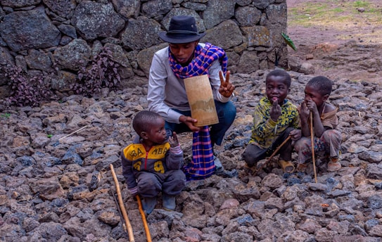 A person in a black hat and checkered scarf kneels on a rocky ground, holding a cylindrical object while gesturing with a peace sign. Two children are standing beside the person with sticks, and one child is sitting on the ground, dressed in a yellow top. The background features a stone wall with some shrubbery.