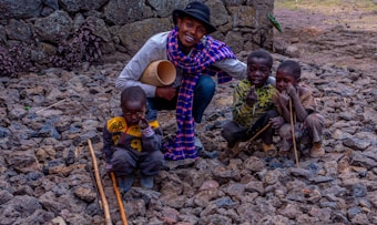A smiling person kneels on rocky ground, surrounded by three children. The person wears a black hat, a colorful blue and purple checkered scarf, and holds a cylindrical object. The children, wearing casual and worn clothing, hold sticks and sit on the rocks. The background features a stone wall and sparse vegetation, giving a rustic and rural impression.