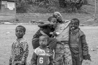Five children are gathered together outdoors, appearing cheerful and bonded. They are dressed in casual, patterned clothing, some with logos and numbers. One child holds what looks like a popsicle, and another has an arm affectionately around a friend. The background shows a dirt ground with a wooden structure and some foliage.