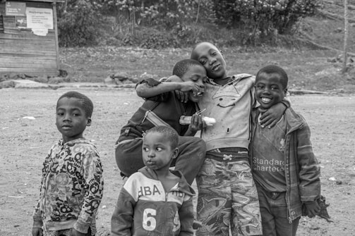 Five children are gathered together outdoors, appearing cheerful and bonded. They are dressed in casual, patterned clothing, some with logos and numbers. One child holds what looks like a popsicle, and another has an arm affectionately around a friend. The background shows a dirt ground with a wooden structure and some foliage.