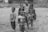 Five children are standing together on a dirt ground, with two of them embracing each other cheerfully. They are dressed in casual clothing and look content. In the background, there is a wooden structure and some vegetation.