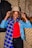 A woman is adjusting a beige hat on her head while wearing a blue and yellow plaid scarf, a red top, a blue denim jacket, and black pants. The background features textured patterned walls and a hanging coat.