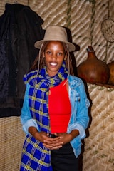 A woman wearing a beige hat, red top, and a blue and yellow checkered scarf stands in front of a woven backdrop. She also wears a denim jacket and has long braided hair. To her left, there is a dark coat hanging, and on the right side, two gourds are placed on a shelf.