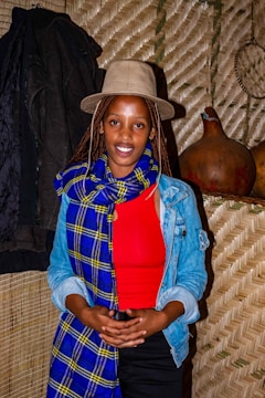 A woman wearing a beige hat, red top, and a blue and yellow checkered scarf stands in front of a woven backdrop. She also wears a denim jacket and has long braided hair. To her left, there is a dark coat hanging, and on the right side, two gourds are placed on a shelf.