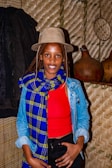 A person wearing a beige hat and a vibrant blue and yellow scarf over a red top and denim jacket is standing indoors against a textured wall. The background includes traditional woven materials and gourds.