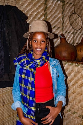 A person wearing a beige hat and a vibrant blue and yellow scarf over a red top and denim jacket is standing indoors against a textured wall. The background includes traditional woven materials and gourds.