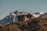 A small wooden cabin sits atop a grassy hill with a backdrop of majestic mountains. The cabin is made of logs and has a rustic appearance. The landscape is serene and the mountains in the distance are capped with snow.