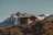 A small wooden cabin sits atop a grassy hill with a backdrop of majestic mountains. The cabin is made of logs and has a rustic appearance. The landscape is serene and the mountains in the distance are capped with snow.