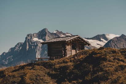 A small wooden cabin sits atop a grassy hill with a backdrop of majestic mountains. The cabin is made of logs and has a rustic appearance. The landscape is serene and the mountains in the distance are capped with snow.