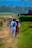 A rural pathway with several people walking along it. The scene includes individuals carrying bundles and walking in groups. The background features lush green fields and distant mountains, with a flag raised on a pole. The atmosphere suggests a peaceful, everyday rural life.