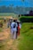 A rural pathway with several people walking along it. The scene includes individuals carrying bundles and walking in groups. The background features lush green fields and distant mountains, with a flag raised on a pole. The atmosphere suggests a peaceful, everyday rural life.
