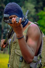 A person is holding a camera equipment, wearing a black balaclava and a green tactical vest. The setting appears to be outdoors with blurred greenery in the background.