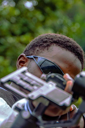 A person wearing dark sunglasses is closely examining a piece of equipment, possibly a camera or a stabilizer. The background is filled with natural greenery, suggesting an outdoor setting.