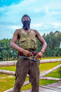 A person wearing a black face mask and an olive green tactical vest stands on a wooden platform outdoors. The background features lush greenery and a blue sky with scattered clouds. The individual is also wearing dark pants and has a waistband with a decorative buckle.