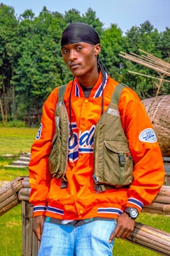 A person wearing an orange jacket over a blue and white shirt, with a black headscarf and a green tactical vest. They are standing outdoors in front of a wooden rail with green trees in the background. The person is also wearing a wristwatch and light blue jeans.