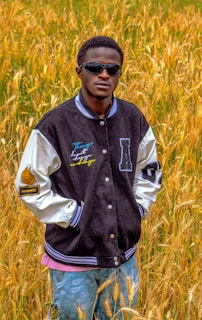 A person wearing sunglasses and a varsity jacket stands in a field of tall, golden grass. The jacket has patches and colorful text on the front. The background features a dense growth of wheat.
