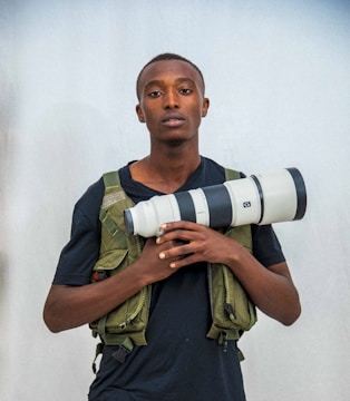 A person stands confidently holding a large professional camera lens. They are wearing a dark shirt and a green tactical vest. The background is a plain light color, and the focus is on the individual and the camera lens they hold.
