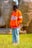 A person wearing an orange jacket with the word 'Florida' written on it, standing in a grassy area. The individual is looking off to the side with hands in pockets. The background features large woven structures and a fence made of bamboo or similar material.
