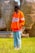 A person wearing an orange jacket with the word 'Florida' written on it, standing in a grassy area. The individual is looking off to the side with hands in pockets. The background features large woven structures and a fence made of bamboo or similar material.