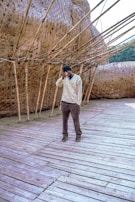 A person stands on a wooden deck next to a large woven bamboo structure. The structure is intricate with interlacing patterns and protruding rods, surrounded by a natural outdoor setting.
