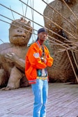 A person wearing a bright orange jacket, blue jeans, and a black headscarf stands confidently on a wooden platform. Behind them are large, woven sculptures resembling animals, made from natural materials like bamboo.