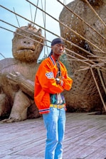 A person wearing a bright orange jacket, blue jeans, and a black headscarf stands confidently on a wooden platform. Behind them are large, woven sculptures resembling animals, made from natural materials like bamboo.