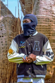 A person wearing a black and white varsity jacket with various letter patches, standing outdoors. Their face is covered with a dark balaclava. The background features large woven structures and tall, thin wooden poles.