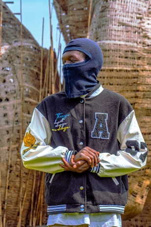 A person wearing a black and white varsity jacket with various letter patches, standing outdoors. Their face is covered with a dark balaclava. The background features large woven structures and tall, thin wooden poles.