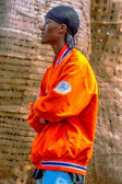 A person stands in profile, wearing an orange jacket with blue and white trim and a patch on the sleeve. They have a black headscarf on and are positioned with arms crossed in front of a textured, woven background.