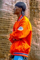 A person stands in profile, wearing an orange jacket with blue and white trim and a patch on the sleeve. They have a black headscarf on and are positioned with arms crossed in front of a textured, woven background.