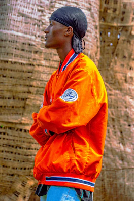 A person stands in profile, wearing an orange jacket with blue and white trim and a patch on the sleeve. They have a black headscarf on and are positioned with arms crossed in front of a textured, woven background.