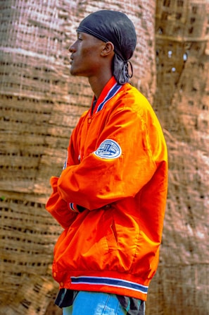 A person stands in profile, wearing an orange jacket with blue and white trim and a patch on the sleeve. They have a black headscarf on and are positioned with arms crossed in front of a textured, woven background.