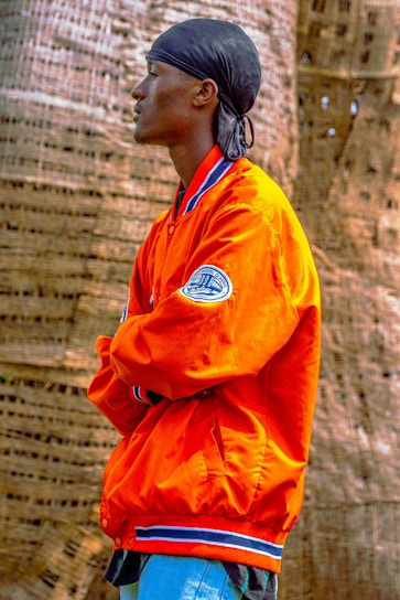 A person stands in profile, wearing an orange jacket with blue and white trim and a patch on the sleeve. They have a black headscarf on and are positioned with arms crossed in front of a textured, woven background.