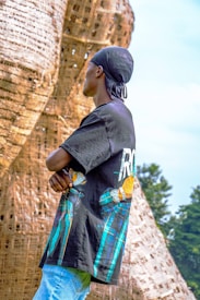 A person wearing a black bandana and a graphic T-shirt stands in front of a large woven structure. The structure appears to be made from natural materials and has a towering, curved design. The T-shirt features colorful graphics and patterns. In the background, there are trees on a clear day with a blue sky.