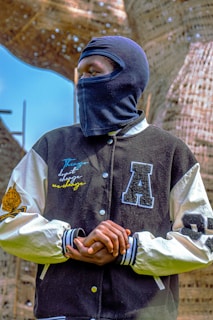 A person wearing a black balaclava and a varsity jacket with white sleeves stands outdoors. The jacket features embroidered text and patches, and the person has their hands clasped together. In the background, there is a structure made of woven material with light filtering through in spots, along with a clear blue sky.