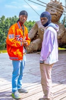 Two young men stand on a wooden platform with a large woven sculpture resembling a giant figure in the background. One wears a bright orange jacket with the number 99 and blue jeans, while the other is in a light-colored hoodie and beige pants. The sky is clear, and there are trees in the background.