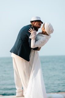 A couple is engaged in a romantic embrace by the sea, with the man leaning slightly forward and the woman in a white outfit looking at him lovingly. Both are smiling, and the background features calm ocean waters and a clear sky.