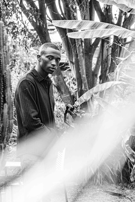 A young man stands in a garden setting, surrounded by plants and trees. He is holding a professional camera and a tripod, indicating a focus on photography or videography. The scene is captured in black and white, giving it a classic, timeless feel. Light flares and shadows add depth to the composition.