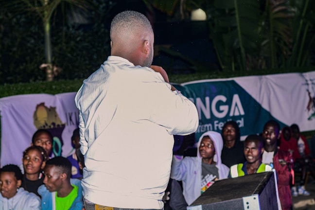 A person wearing a white jacket is standing in front of an audience. The background shows a banner with text partially visible. The event seems to be taking place outdoors at night. The audience consists of various people, some wearing hoodies and jackets.
