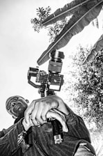 A person is holding a camera with a gimbal, captured from a low angle. The background features large leaves and foliage, creating a natural setting. The image is in black and white.