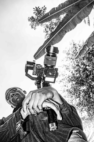 A person is holding a camera with a gimbal, captured from a low angle. The background features large leaves and foliage, creating a natural setting. The image is in black and white.