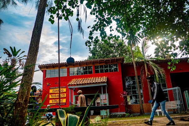 A vibrant outdoor scene with a red building featuring large windows and a brown tiled roof. The signage on the building reads 'Isange Paradise Resort'. The area is surrounded by lush greenery, including trees and various plants. Two people are walking in the foreground, and a swing set is partially visible.