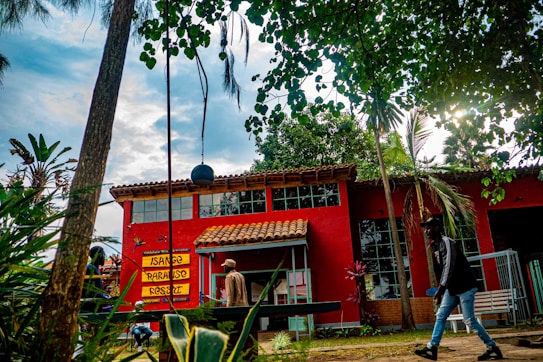 A vibrant outdoor scene with a red building featuring large windows and a brown tiled roof. The signage on the building reads 'Isange Paradise Resort'. The area is surrounded by lush greenery, including trees and various plants. Two people are walking in the foreground, and a swing set is partially visible.