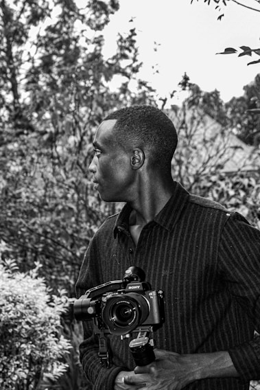 A man stands outdoors holding a camera, with his head turned to the side. He is surrounded by dense foliage and trees in the background, creating a natural setting. The image is black and white, emphasizing contrasts and textures.