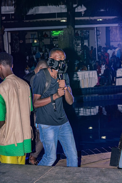 A person holds a stabilizing gimbal while recording with a camera. The setting appears to be a bustling indoor venue or event, possibly a party or gathering, with people seated in the background. The lighting is dim with some artificial lights creating a contrast.