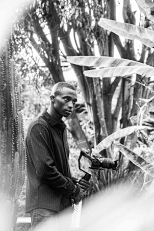 A black and white photo captures a person standing amidst a lush, leafy environment. The individual holds a camera mounted on a stabilizer, appearing focused and contemplative. Large leaves and tall trees provide a dense backdrop, suggesting a setting rich with foliage.