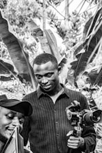 A black and white photo captures two men, one holding a camera with a stabilizer, and the other smiling while looking at a tablet or phone. They are outdoors, surrounded by large leaves from tropical plants.